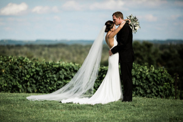 Bride with long veil and groom embracing in front of grape vines.