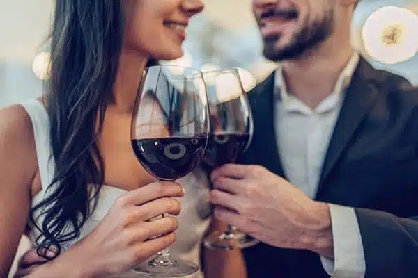 Inn 2 A couple dressed in formal attire clink glasses of red wine. They smile at each other, creating a warm and intimate atmosphere. The background is softly blurred with glowing lights, suggesting a romantic or celebratory setting.