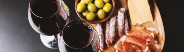 Two glasses of red wine with meat, cheese, and olives in the background.